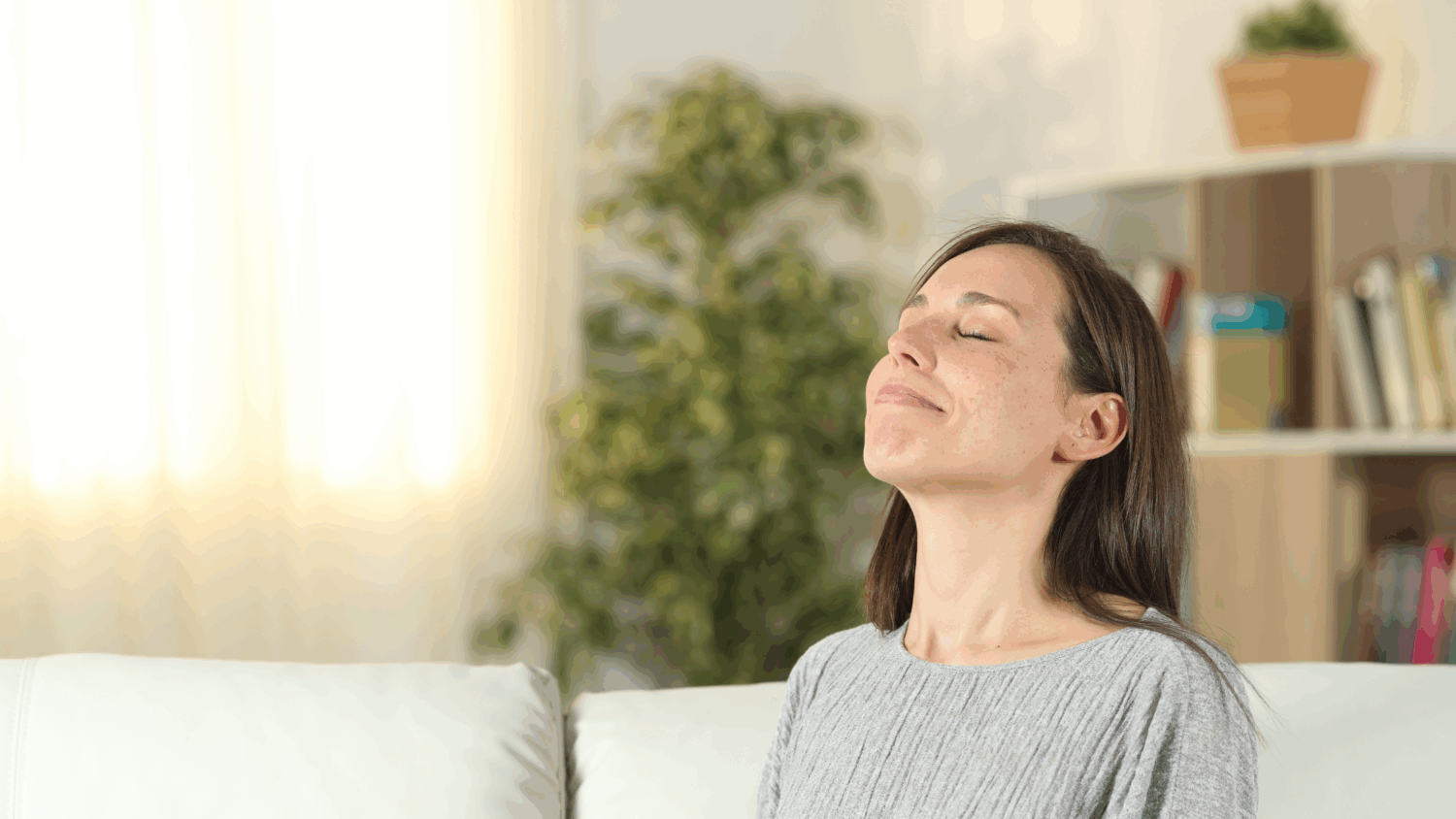 A smiling woman sits on a sofa, breathing deeply to enjoy fresh, clean air in a bright living room.
